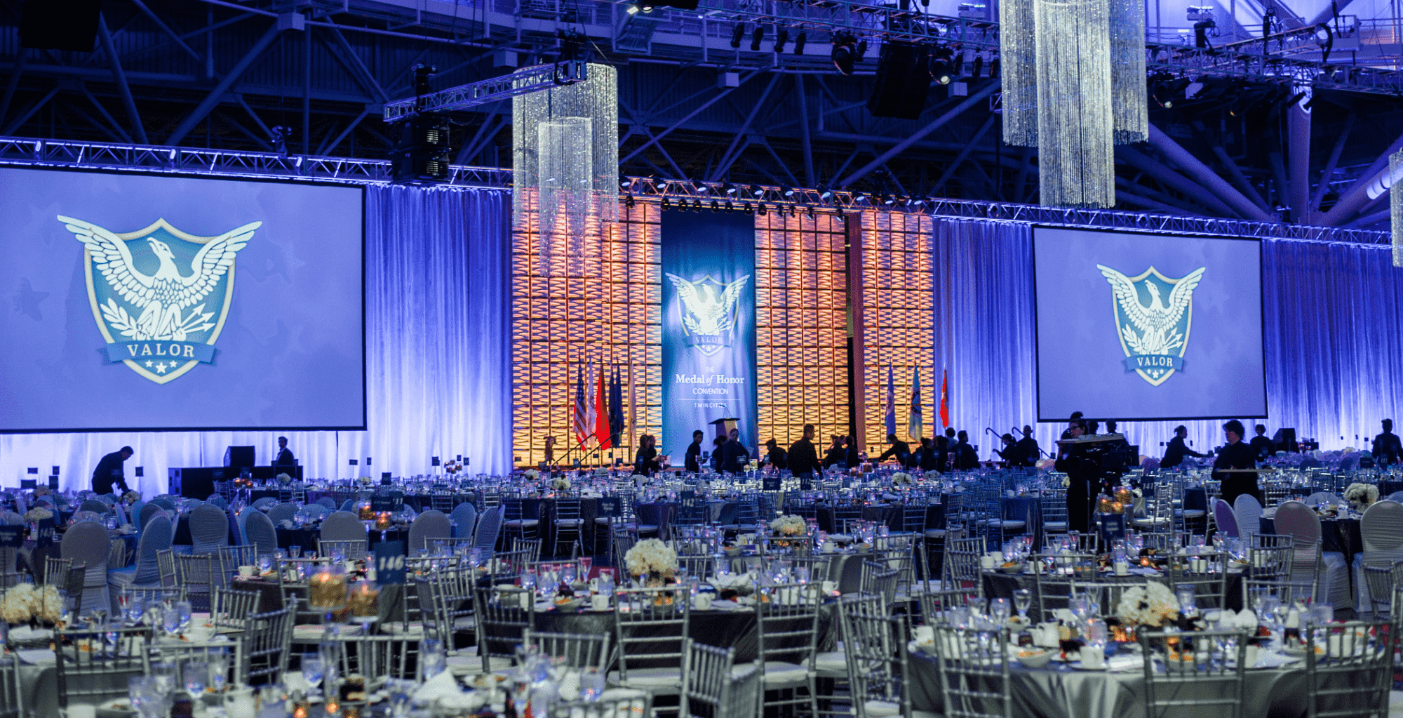 A ballroom filled decorated tables with flowers and silver tablecloths. Blue and orange colored lights on stage backdrops. Flags on the stages and company logo projected on screens and stage backdrop.