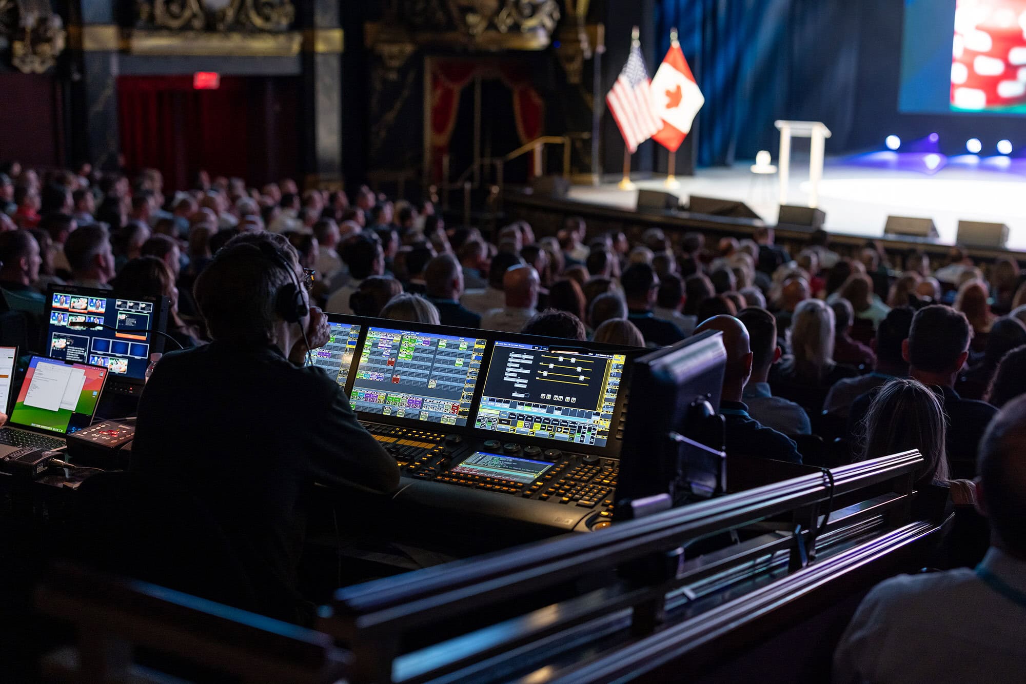 PB1A9913 - metroConnections A producer sits at a desk with many laptops, looking at the stage thoughtfully. The audience is full of people paying rapt attention.