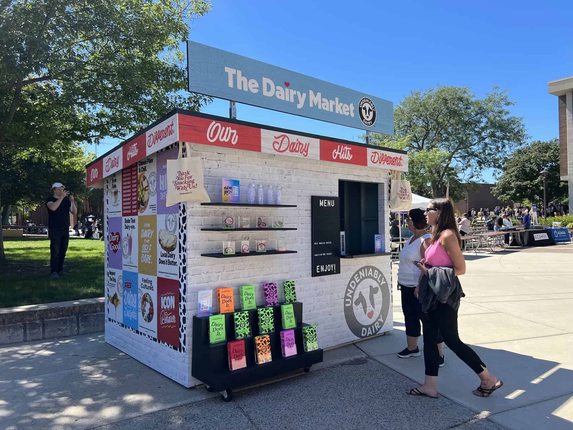 Midwest Dairy Custom Build - metroConnections A small fabricated building called The Dairy Market. There are shelves for literature and swag, and a window where attendees can order off a menu.