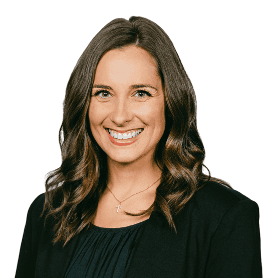 Professional headshot of a smiling woman with long brown hair, wearing a black blazer and gold cross necklace.