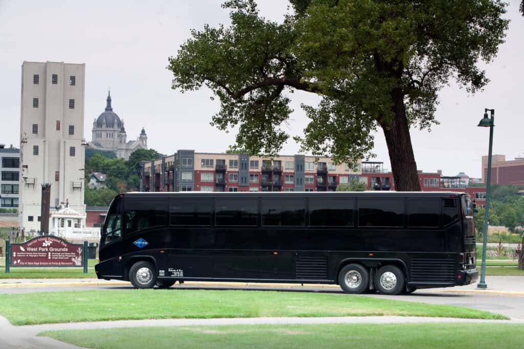 Executive Motorcoach - metroConnections Black tour bus parked on a city street with a grassy area, large tree, and modern buildings in the background at dusk/dawn lightettings are neutral in color.