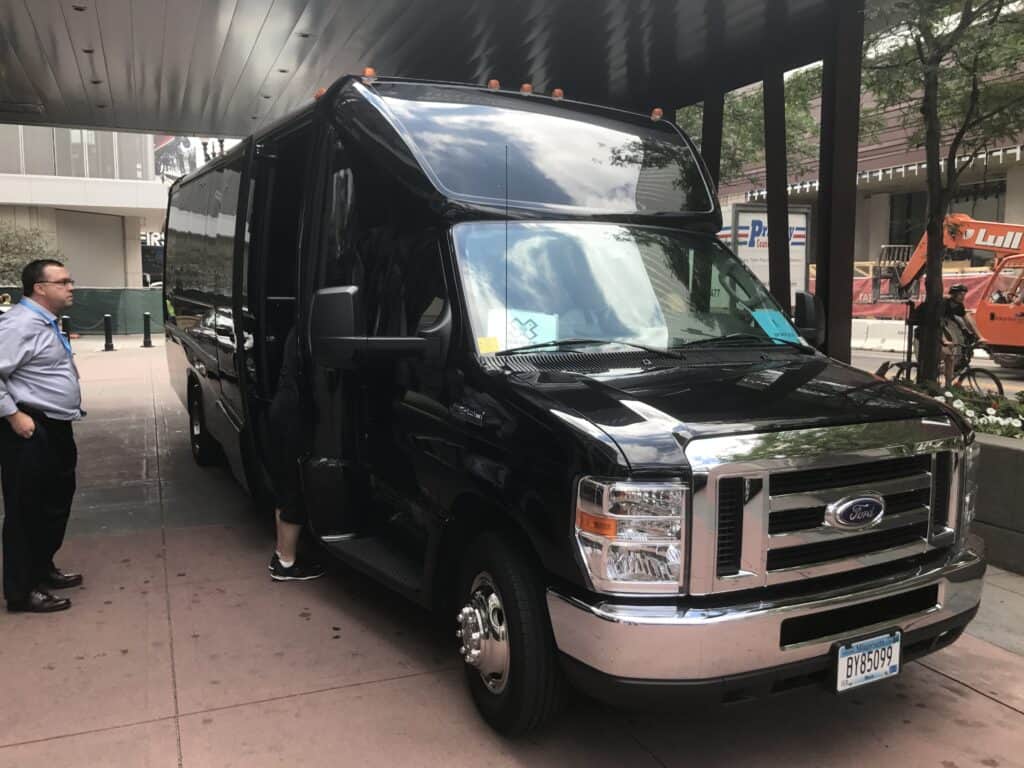 IMG_1516 - metroConnections Front view of a large black Ford shuttle/van with orange roof clearance lights, parked by a curb while a man in a blue shirt stands nearby.