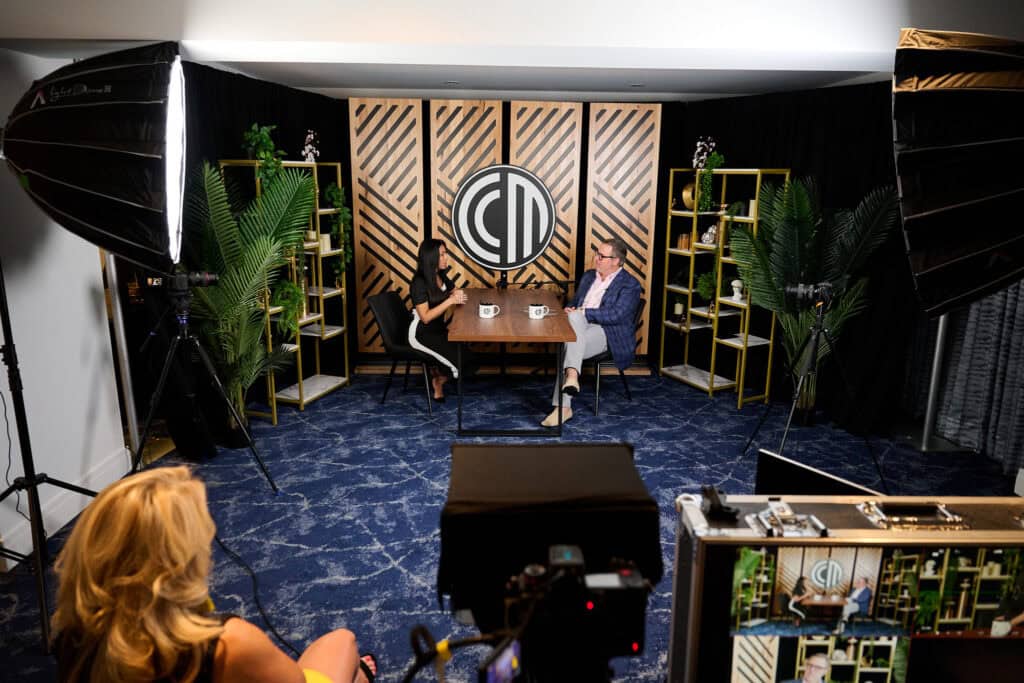 CCM - metroConnections Two people sit across from each other at a wooden table in a studio set, with cameras and studio lights filming a conversation against a patterned backdrop and logo center stage.
